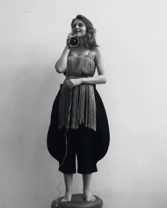 Monochrome portrait of a woman wearing an avant-garde fringe top and black culottes, holding an old telephone to her ear in studio lighting.