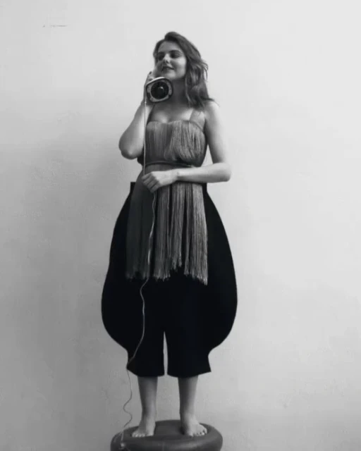 Monochrome portrait of a woman wearing an avant-garde fringe top and black culottes, holding an old telephone to her ear in studio lighting.