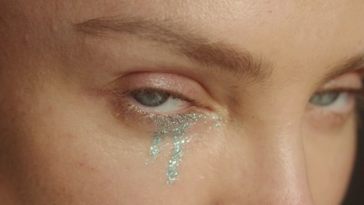 Close-up of a woman's face with grey eyes and silver glitter tears running down her cheek, set against a dark background.