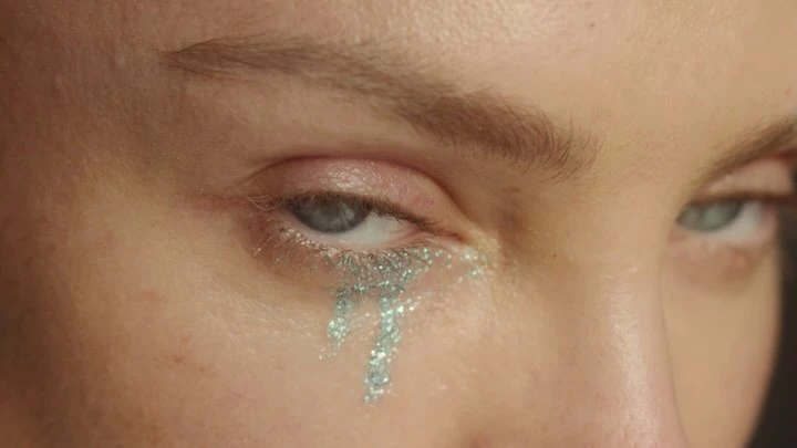 Close-up of a woman's face with grey eyes and silver glitter tears running down her cheek, set against a dark background.