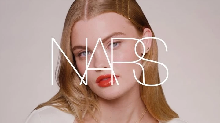 Portrait of a fair-skinned model with blonde hair, coral lips, and bobby pins, overlaid with the NARS brand logo in a beauty context.