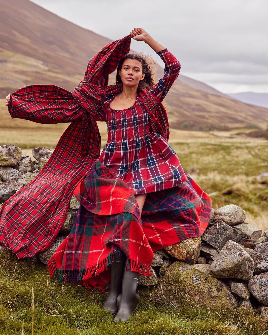 A woman in a long, red plaid dress stands on a stone wall in a field with mountains in the background, holding a scarf.