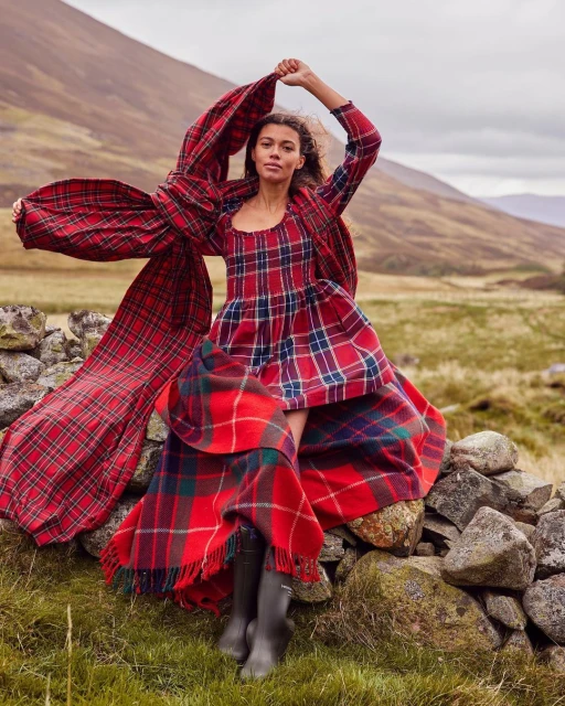 A woman in a long, red plaid dress stands on a stone wall in a field with mountains in the background, holding a scarf.
