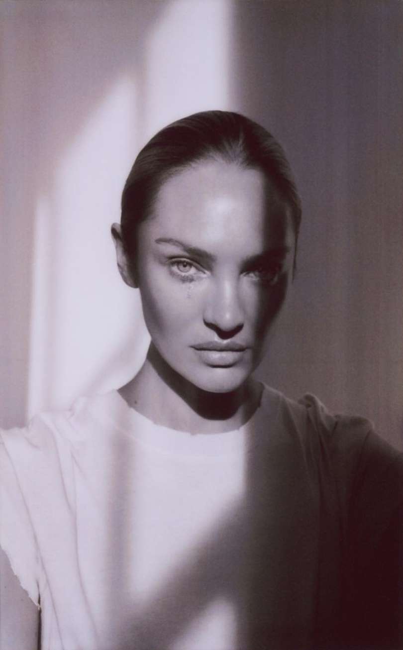 Close-up portrait of a woman with neutral expression in a white shirt, dramatic side lighting, and slicked-back hair, on a neutral background.