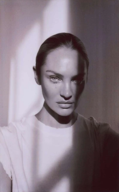 Close-up portrait of a woman with neutral expression in a white shirt, dramatic side lighting, and slicked-back hair, on a neutral background.