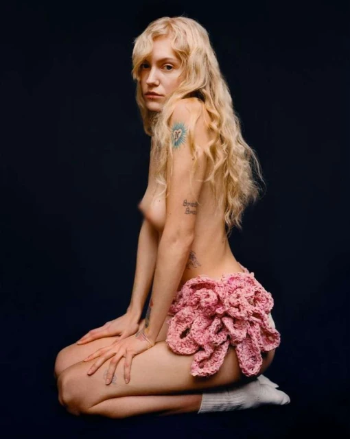 Blonde-haired woman poses kneeling in a photo studio. She has tattoos and is wearing a pink skirt and socks against a dark background.