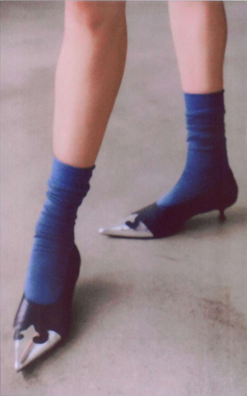 Woman wearing blue socks and black, silver pointed-toe heeled shoes standing on a grey surface in a fashion shot.