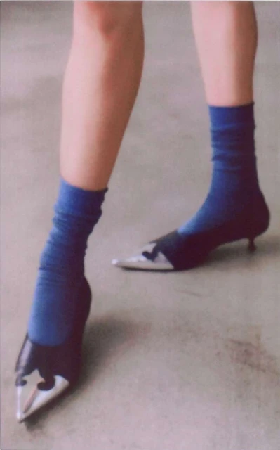 Woman wearing blue socks and black, silver pointed-toe heeled shoes standing on a grey surface in a fashion shot.