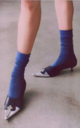 Woman wearing blue socks and black, silver pointed-toe heeled shoes standing on a grey surface in a fashion shot.