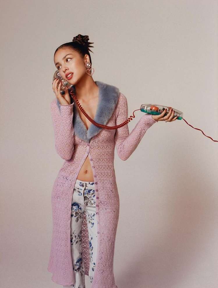 Olivia Rodrigo poses in a long pink cardigan, floral pants and holds a transparent telephone in a bright, minimalist photo studio.