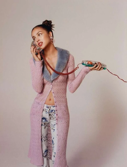 Olivia Rodrigo poses in a long pink cardigan, floral pants and holds a transparent telephone in a bright, minimalist photo studio.