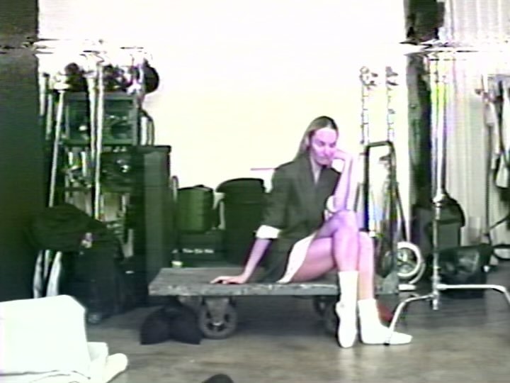 Adult woman sits on a rolling platform in a photo studio, wearing a blazer with a white dress and boots, surrounded by equipment.