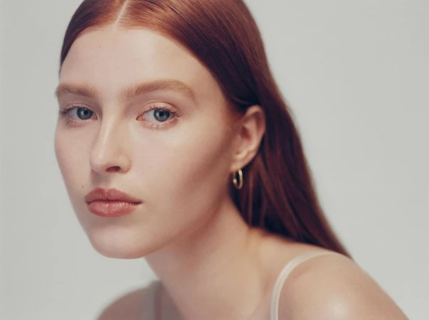 Close-up portrait of a woman with red hair and hoop earrings against a neutral background by Heather Hazzan