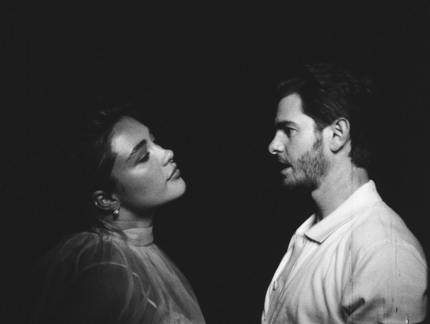 Black and white portrait of a woman in a sheer dress and a man in a collared shirt, looking at each other against a dark background.