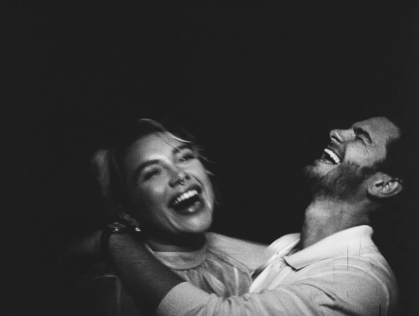 A black and white photograph features a woman and man laughing joyfully in an embrace, set against a dark background with candid and emotional tones.
