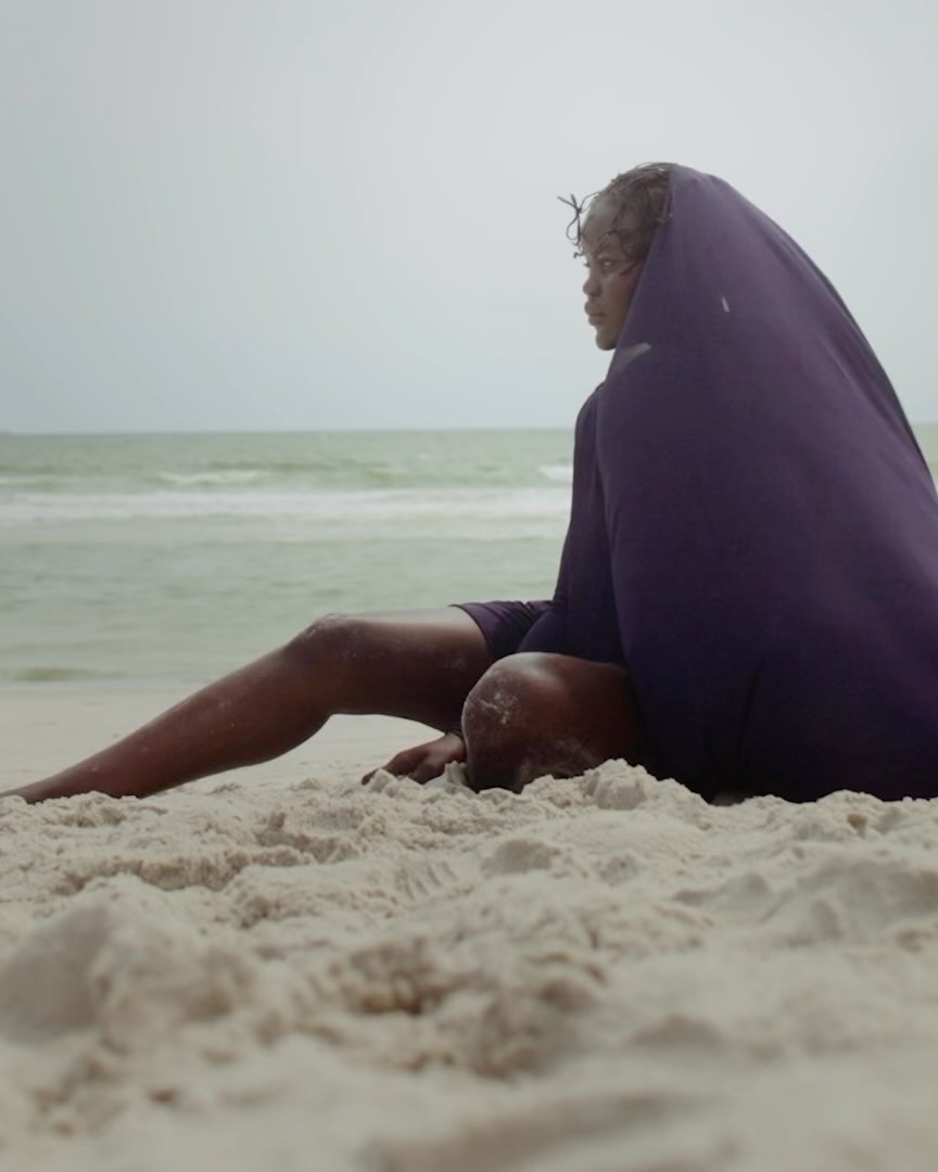 Woman sitting on a sandy beach covered with a purple sheet, ocean and cloudy sky in the background, contemplative pose.