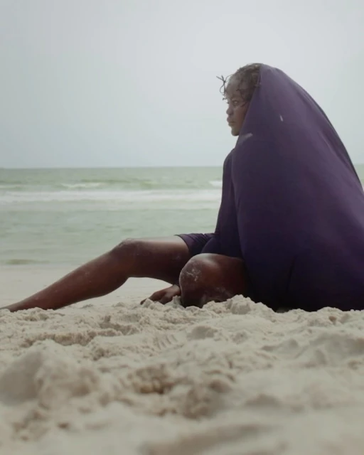 Woman sitting on a sandy beach covered with a purple sheet, ocean and cloudy sky in the background, contemplative pose.