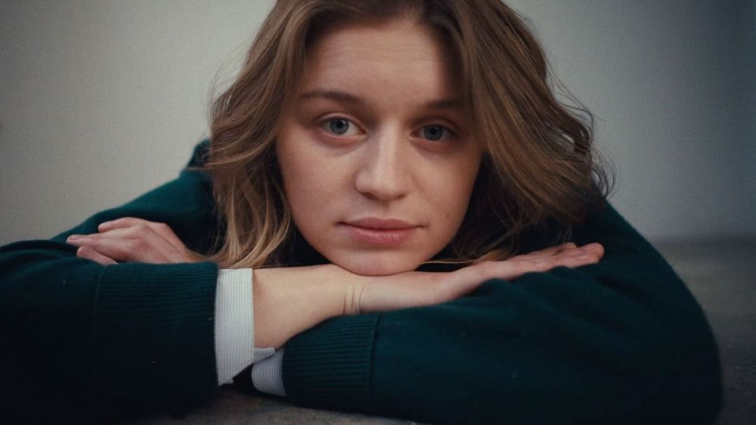 Close-up portrait of a young adult female with fair skin, resting her chin on her hands, wearing a green sweater indoors.
