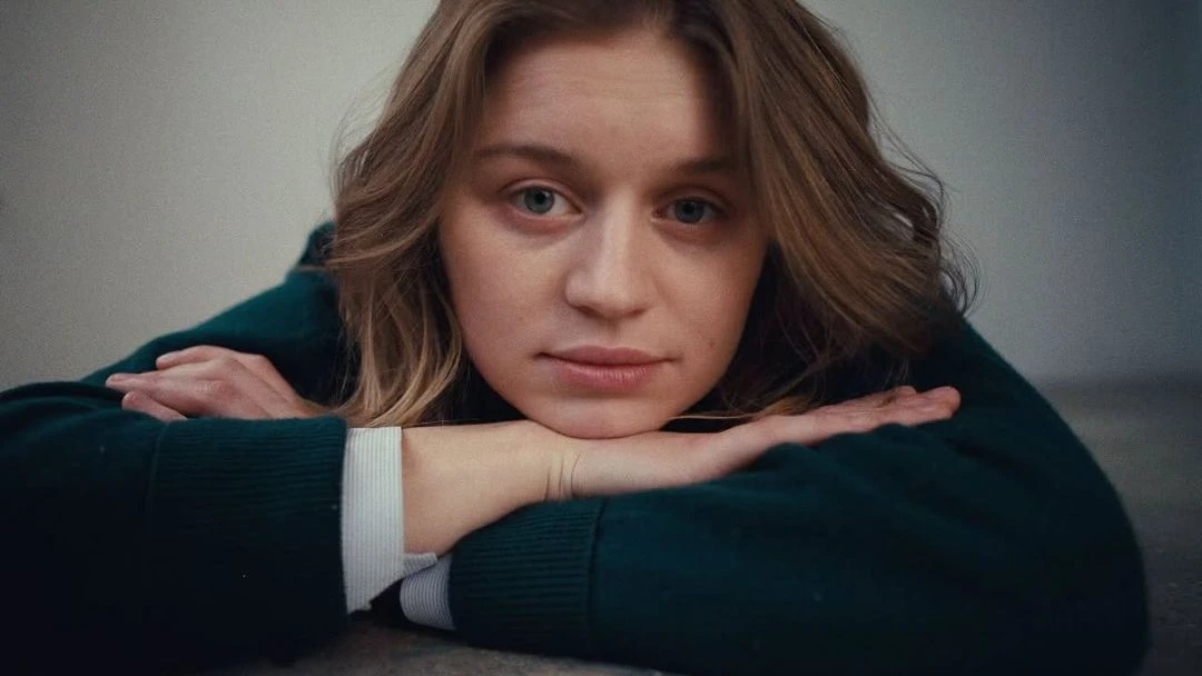 Close-up portrait of a young adult female with fair skin, resting her chin on her hands, wearing a green sweater indoors.