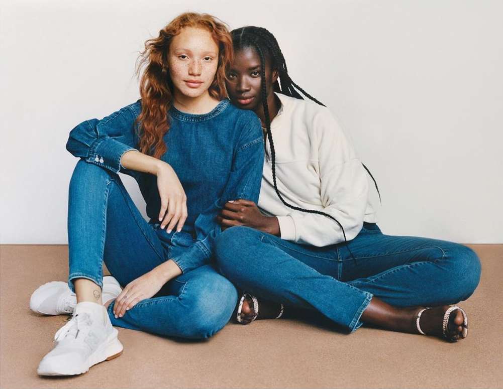 Two women, one with red hair and the other with black braided hair, sit close together indoors wearing blue jeans and light-colored tops.