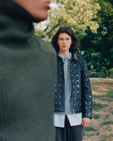 Two young male fashion models pose in a park setting, one wearing a green ribbed turtleneck, the other wearing a black quilted jacket.