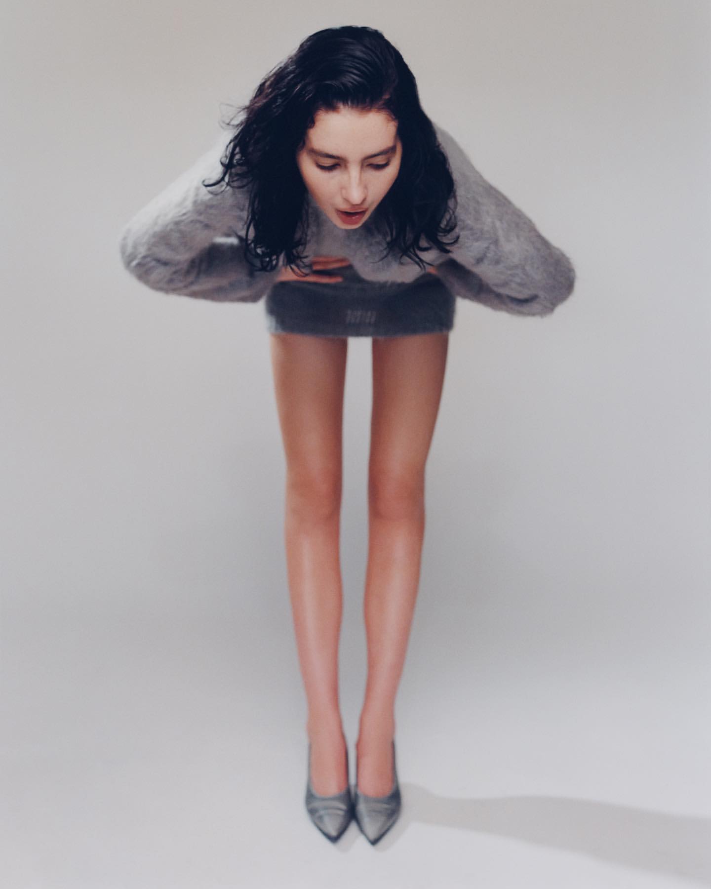 Model with dark wet-look hair bending forward in a grey knit sweater and matching skirt against a white background