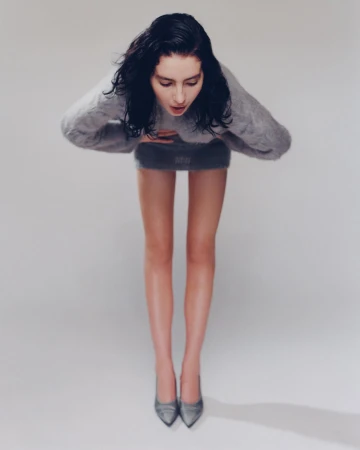 Model with dark wet-look hair bending forward in a grey knit sweater and matching skirt against a white background