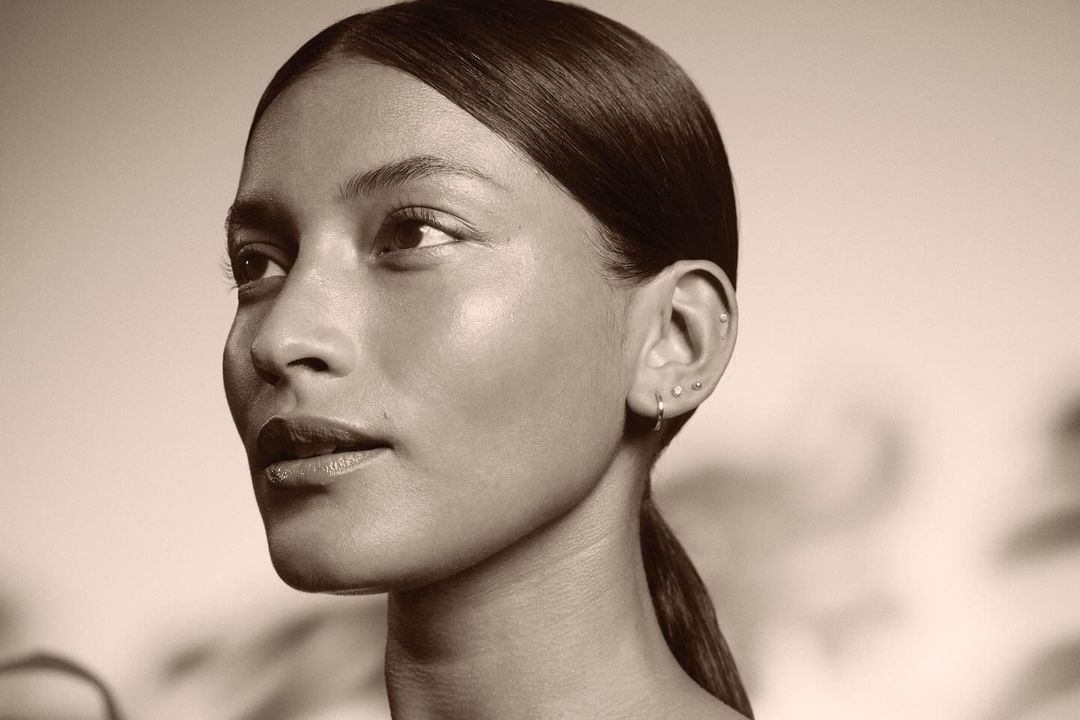 Close-up portrait of a woman with sleek dark hair pulled back, multiple ear piercings, minimal makeup, and soft neutral lighting.