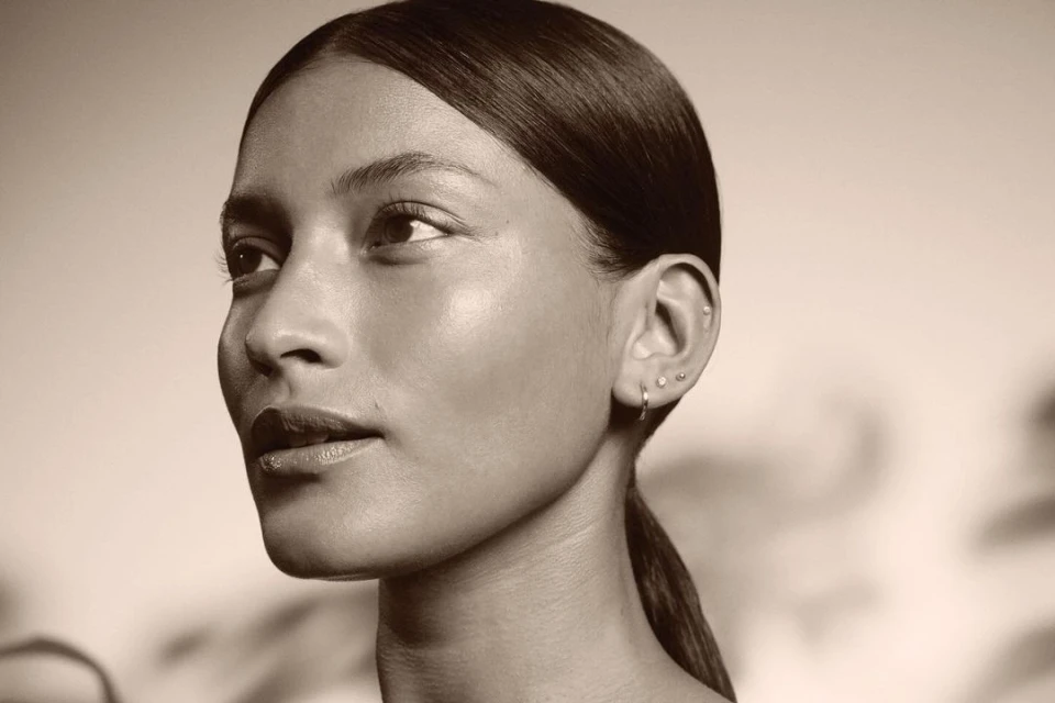 Close-up portrait of a woman with sleek dark hair pulled back, multiple ear piercings, minimal makeup, and soft neutral lighting.