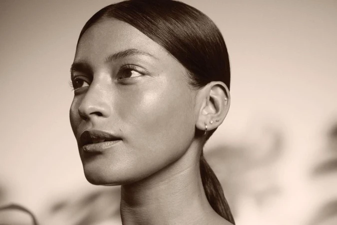 Close-up portrait of a woman with sleek dark hair pulled back, multiple ear piercings, minimal makeup, and soft neutral lighting.