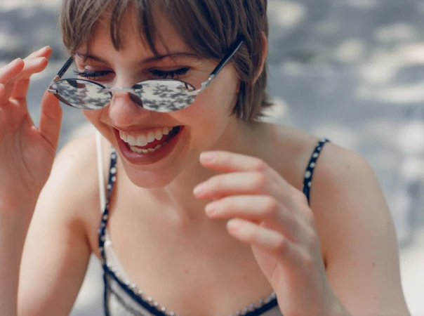 Close-up portrait of a smiling woman with short brown hair wearing small silver sunglasses and a polka dot top outdoors.