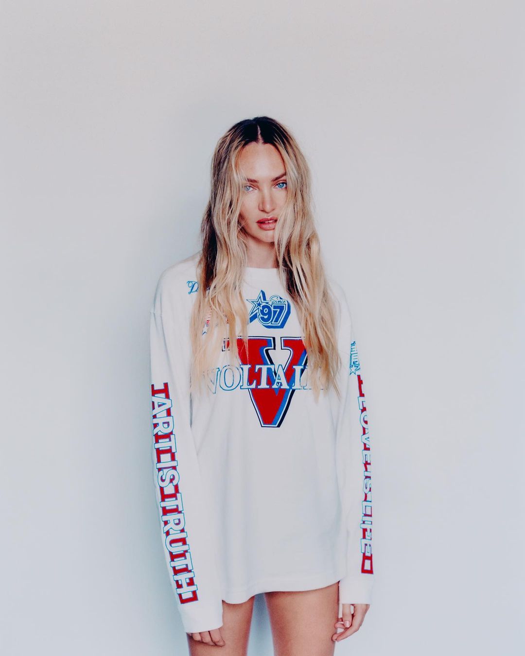 Blonde female fashion model poses in a white, long-sleeved graphic tee against a white backdrop in a medium studio shot.