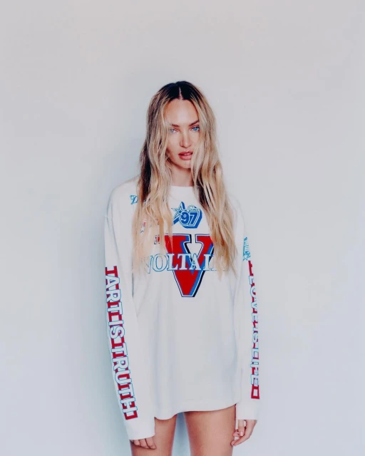 Blonde female fashion model poses in a white, long-sleeved graphic tee against a white backdrop in a medium studio shot.