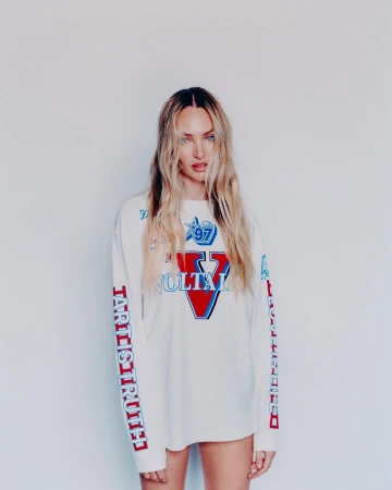 Blonde female fashion model poses in a white, long-sleeved graphic tee against a white backdrop in a medium studio shot.