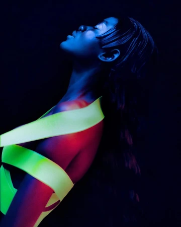 Portrait of a woman with eyes closed, face angled upwards, wrapped in a neon yellow ribbon against a black background with blue lighting.