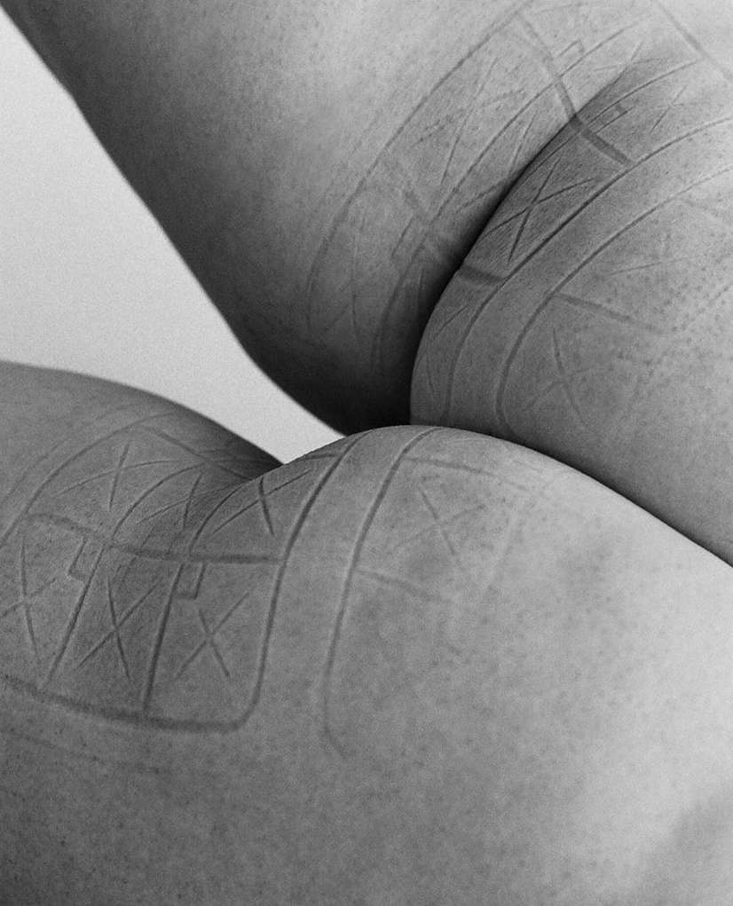 Close-up studio shot of human legs with geometric patterns inscribed on skin in monochrome. Focus on skin texture, minimalist aesthetic.