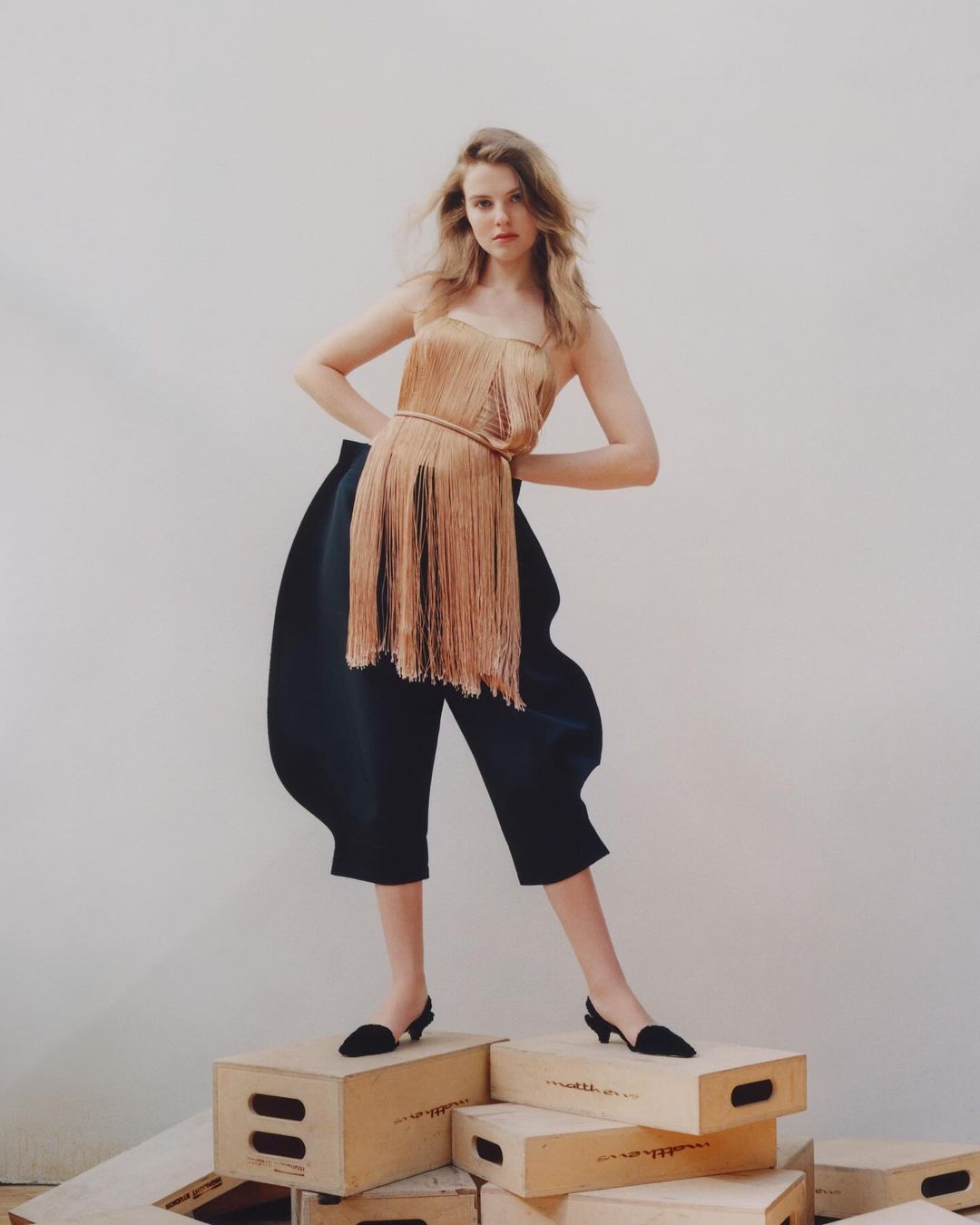 Full-length shot of a young woman in a gold fringe top and dark blue culottes posing on wooden boxes in a bright, white-walled studio.