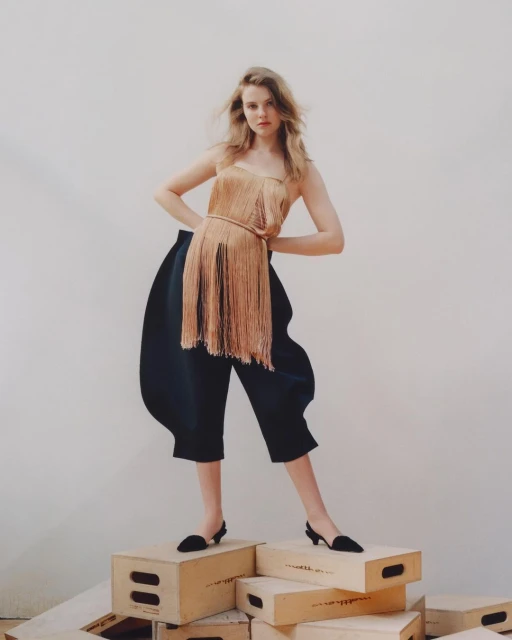 Full-length shot of a young woman in a gold fringe top and dark blue culottes posing on wooden boxes in a bright, white-walled studio.