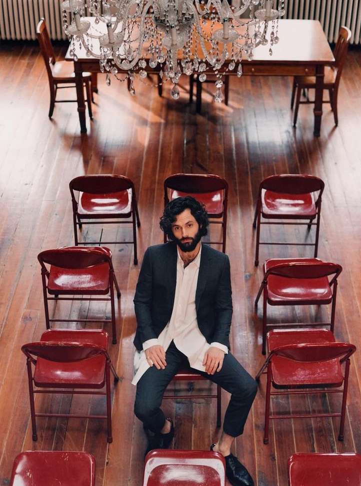Man in a suit seated among red folding chairs under a chandelier in a room with wooden floors and a table with chairs.