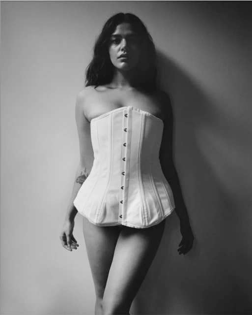 Monochrome full shot of a woman wearing a corset top against a neutral wall, showcasing fashion and posing for the camera.