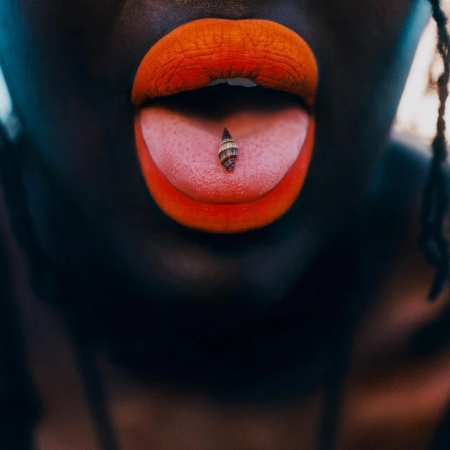 Close-up of an African American adult female with a seashell on her tongue, wearing vibrant orange lipstick, and dark skin, against a dark background.