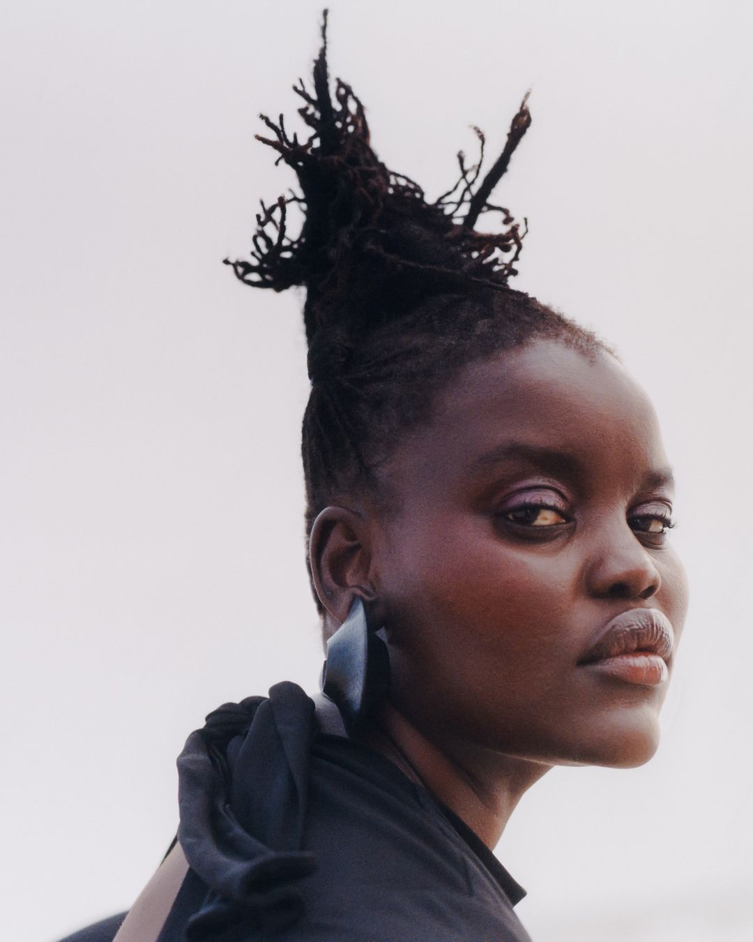 Portrait of a Black woman with dark hair styled in a high updo, wearing dark clothing and a large earring against a light background.