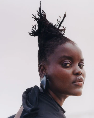 Portrait of a Black woman with dark hair styled in a high updo, wearing dark clothing and a large earring against a light background.
