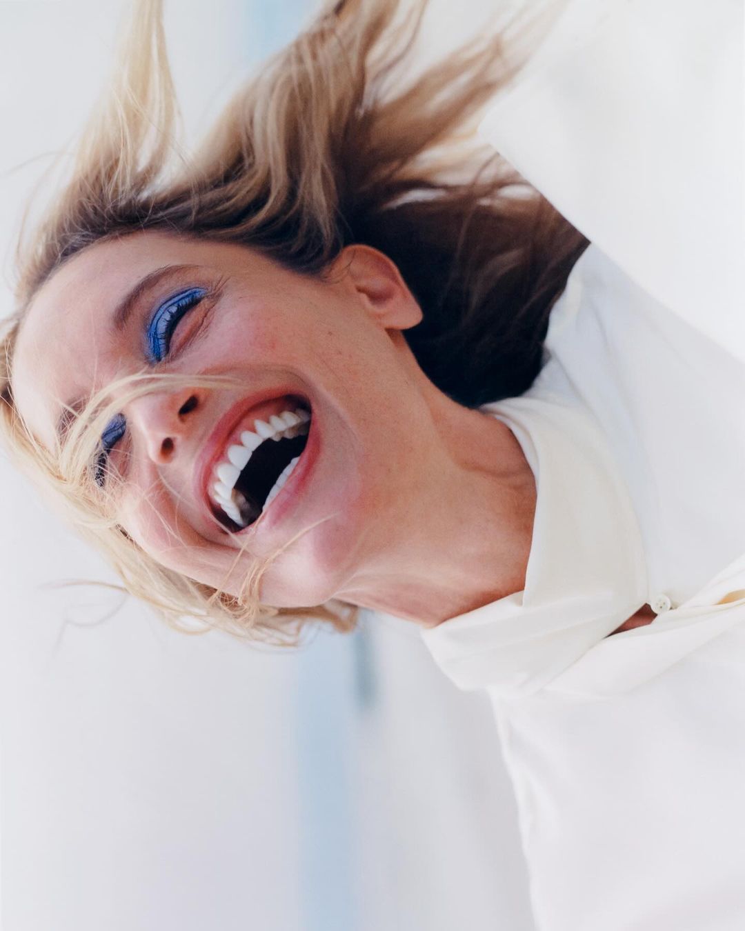 Portrait of a laughing adult female with blond hair and blue eyeshadow wearing a white top, captured with a bright, upward perspective.