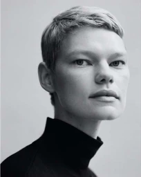 Close up monochrome portrait of a woman with short blonde hair wearing a black turtleneck shirt against a neutral background.