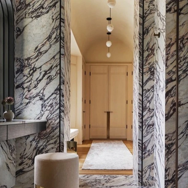 Hallway featuring marble walls and flooring, leading to white doors with a light beige rug. Decorative lighting overhead.