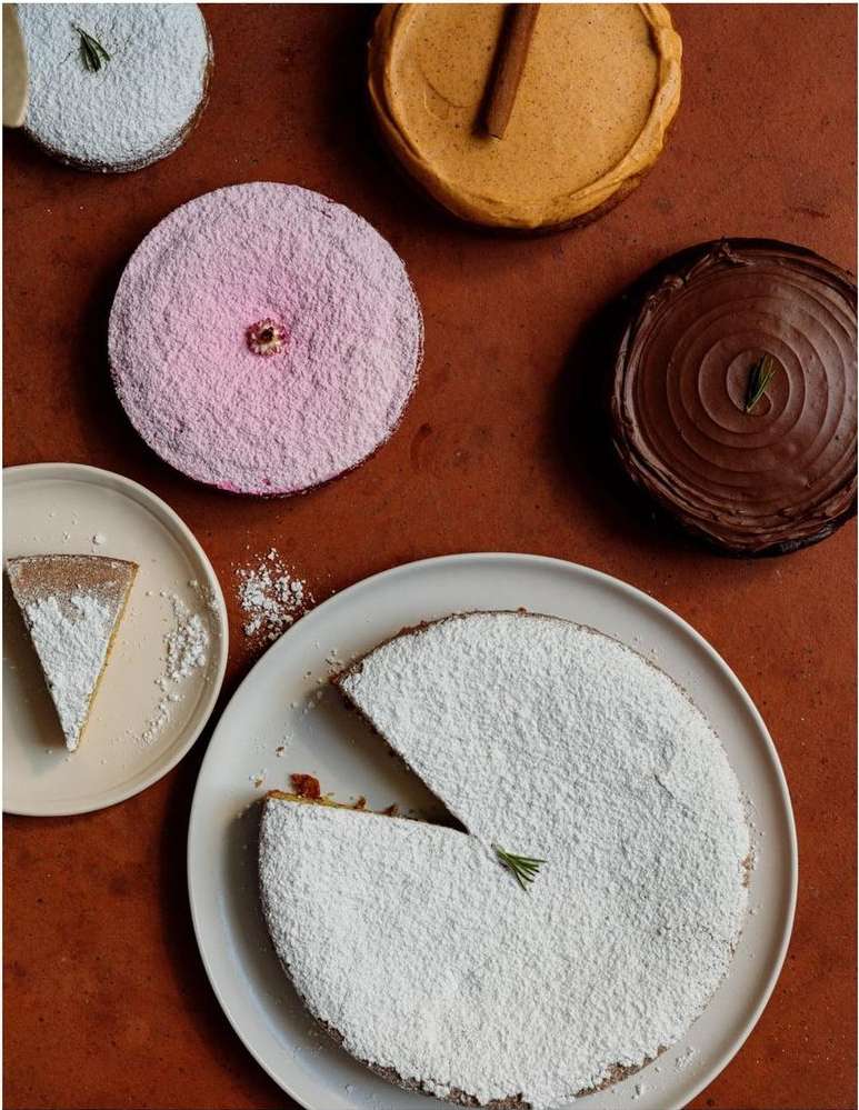 Overhead view of multiple cakes with colorful toppings and powdered sugar on a brown surface, food photography, top-down perspective.