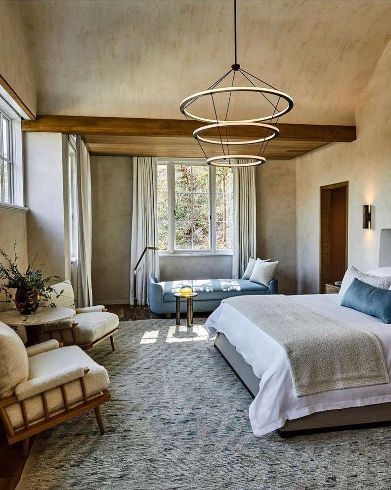 Bedroom interior features a bed with blue pillows, two chairs, a daybed, and a hanging circular chandelier, creating a serene and stylish space.
