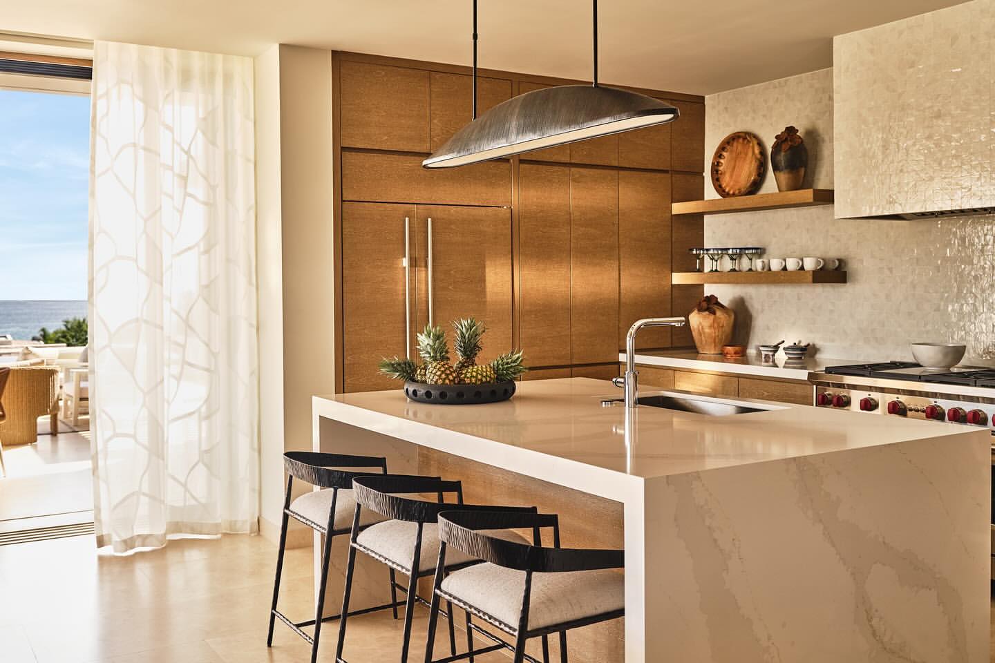 Bright and airy home kitchen featuring a white countertop island with modern black bar stools, hardwood cabinets, and ocean views.