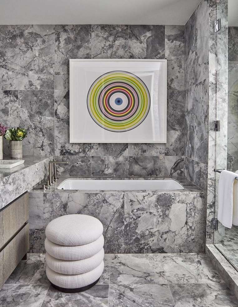 Modern bathroom interior with gray marble walls, a freestanding white tub, round upholstered stool, and framed abstract art in soft light.